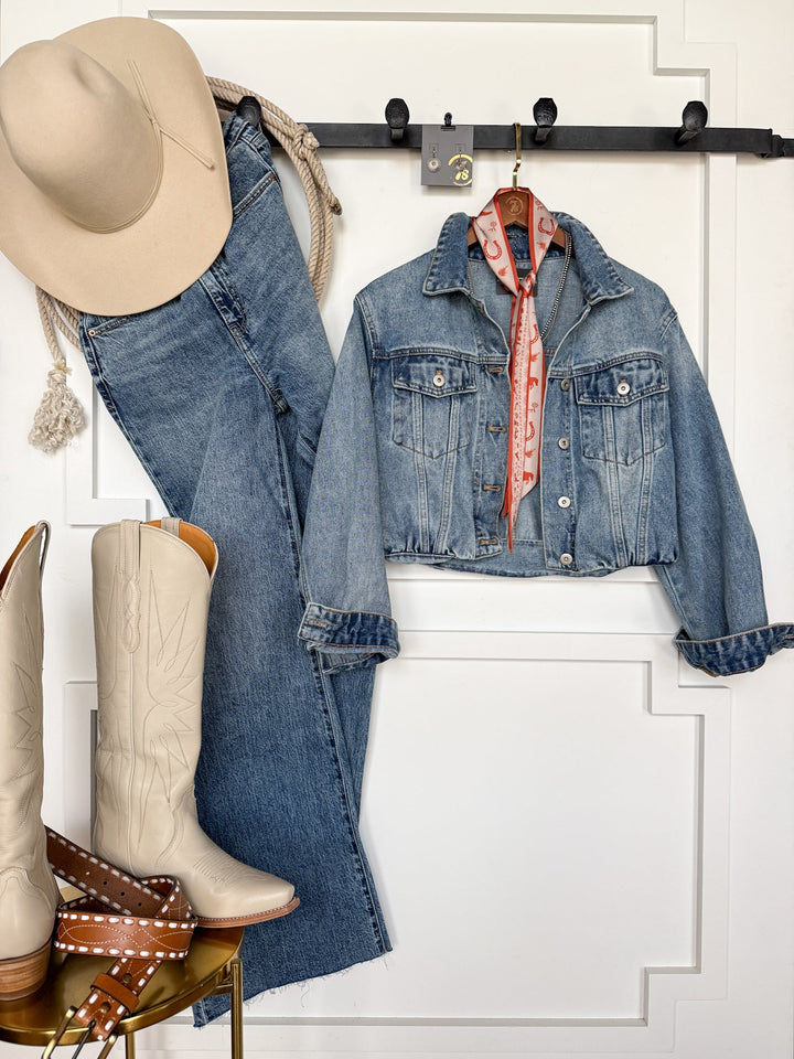 A beige cowboy hat, rope, medium wash denim jeans, the Memphis Denim Jacket by Savannah Sevens western life{&}style, a red bandana, cream cowboy boots, and a brown studded belt are arranged neatly against a white wall.