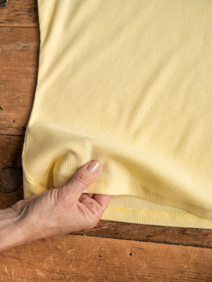A hand gently pinches the hem of the light yellow Merrill Butter Modal Tank by Savannah Sevens western life{&}style, displayed flat on a wooden surface.