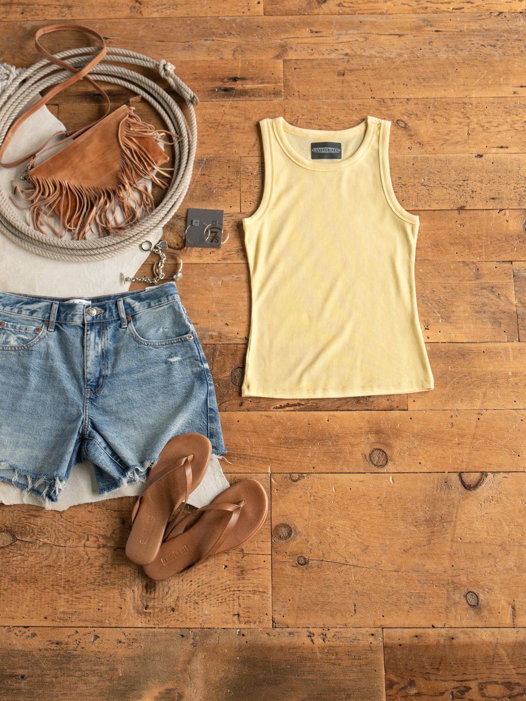 Flat lay of a casual summer outfit: Savannah Sevens western life{&}style Merrill Butter Modal Tank, denim shorts, brown fringe bag, tan sandals, belt, and rope accessory arranged on a wooden floor.