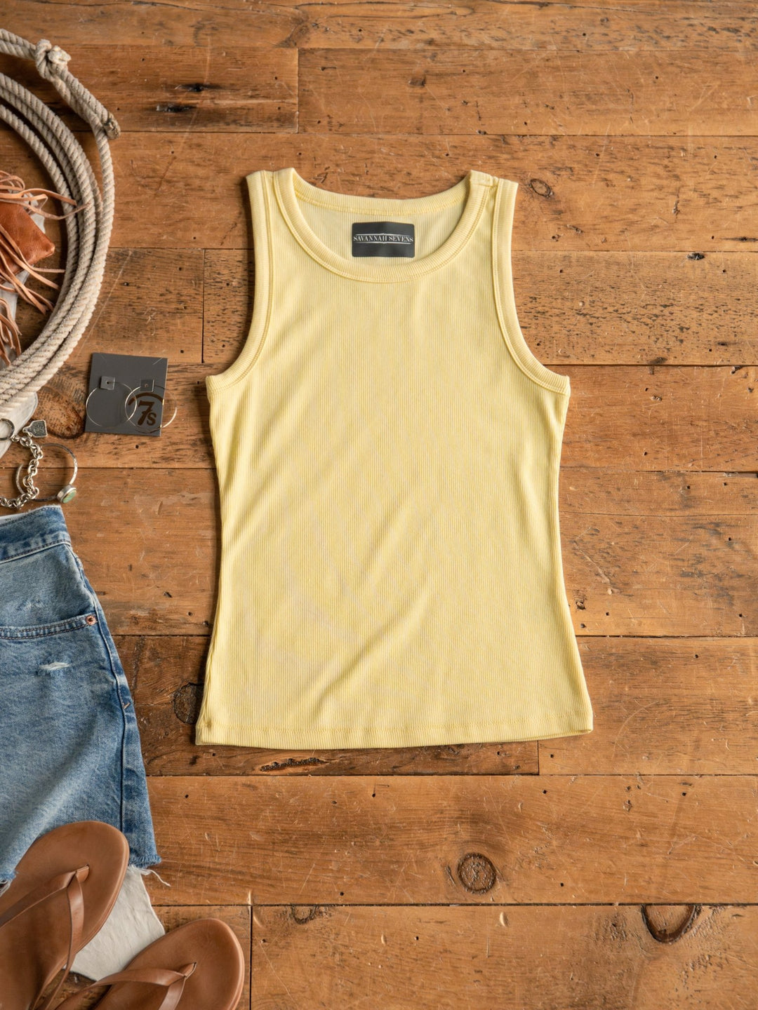 The Merrill Butter Modal Tank by Savannah Sevens western life{&}style is laid flat on a wooden floor beside denim shorts with a price tag, tan sandals, a thick rope, and a brown hat.