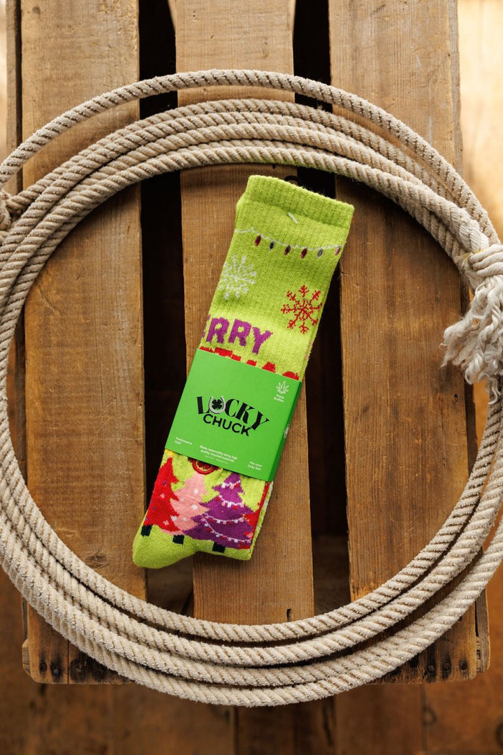 A pair of Merry Christmas Y’all Crew Socks {Green} by Savannah Sevens western life{&}style, featuring festive patterns and a bright green hue, sits on a wooden surface encircled by a coiled white rope.