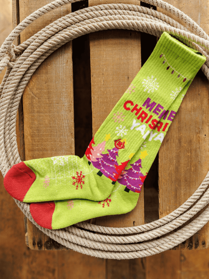 A pair of Merry Christmas Y’all Crew Socks (Green) by Savannah Sevens western life{&}style, featuring red toes, snowflakes, colorful Christmas trees, and "MERRY CHRISTMAS," displayed inside a coiled rope on wood.