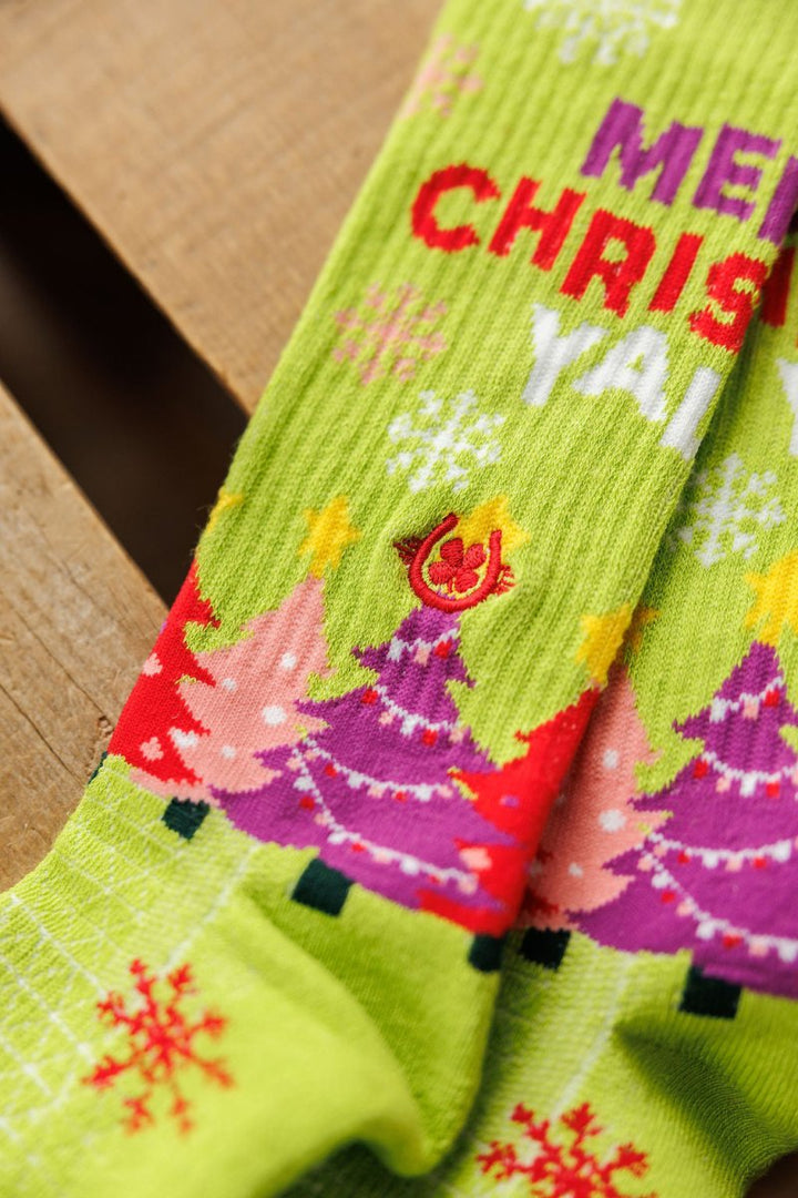 Close-up of Savannah Sevens western life{&}style Merry Christmas Y’all Crew Socks {Green}, featuring colorful Christmas trees, snowflakes, and festive text. Breathable hemp socks with odor resistance rest on a wooden surface.