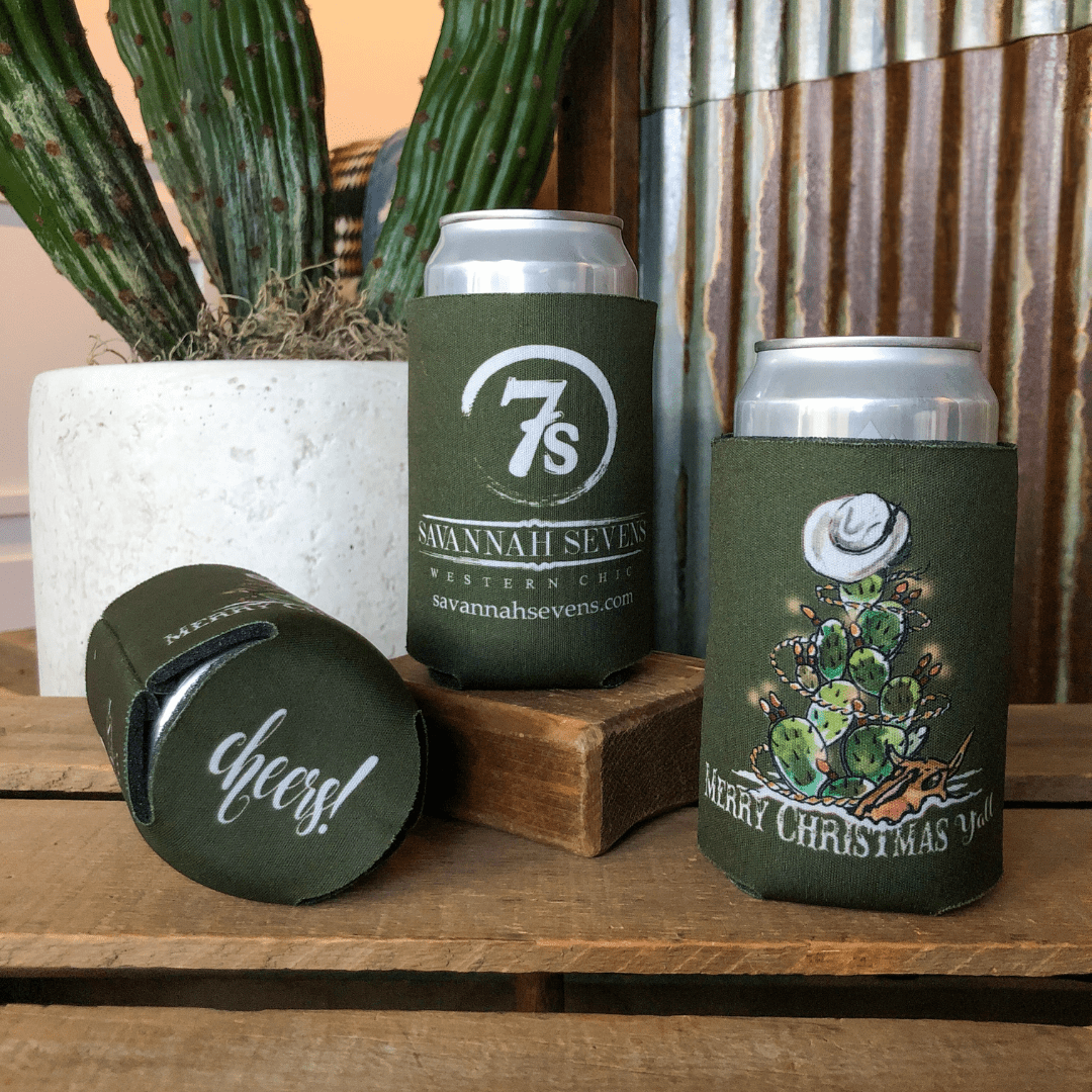 Three canned drinks in dark green koozies, including the "Merry Christmas Y'all Koozie" by Savannah Sevens western life{&}style, sit on a wooden surface with a potted cactus in the background.