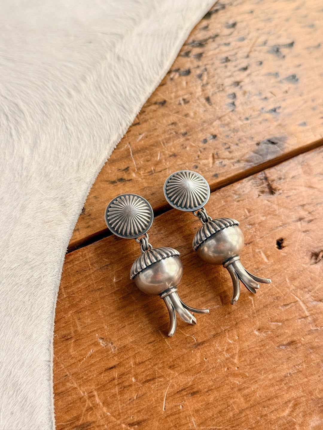 The Mesa Earrings by Savannah Sevens western life{&}style feature sterling silver sunburst studs with dangling pomegranate-shaped charms, displayed on wood beside light fur. These handmade squash blossom earrings are made in the USA.
