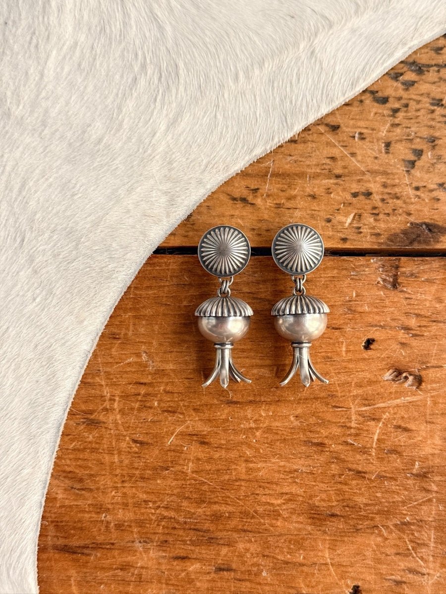 The Mesa Earrings by Savannah Sevens western life{&}style, featuring sterling silver round textured studs and drop-shaped pendants, rest on a wooden surface next to white cowhide.