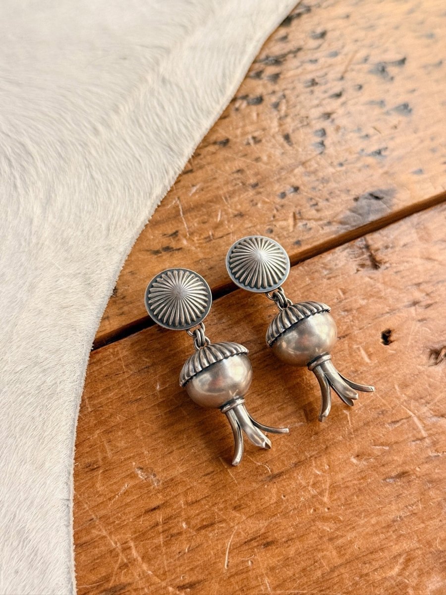 The Mesa Earrings by Savannah Sevens western life{&}style feature sterling silver sunburst studs with dangling pomegranate-shaped charms, displayed on wood beside light fur. These handmade squash blossom earrings are made in the USA.