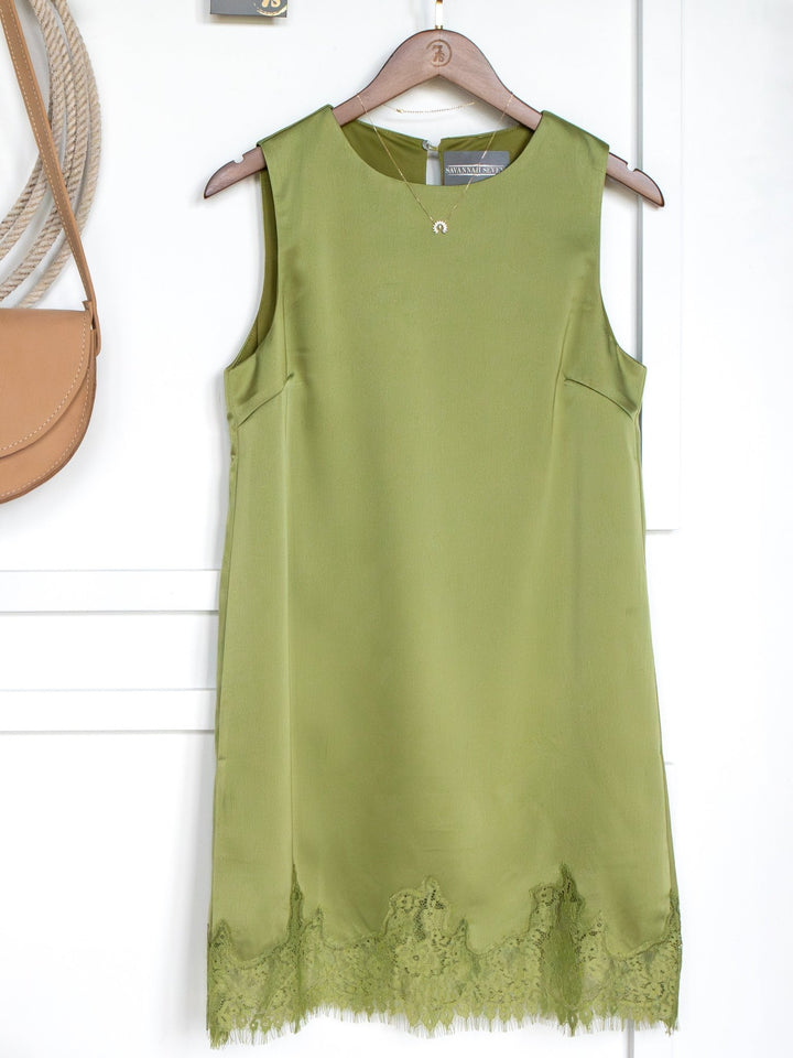 The Mesena Light Olive Satin Dress by Savannah Sevens western life{&}style, featuring a sleeveless design with a lace hem, hangs on a wooden hanger against a white wall. A tan shoulder bag peeks in from the left.