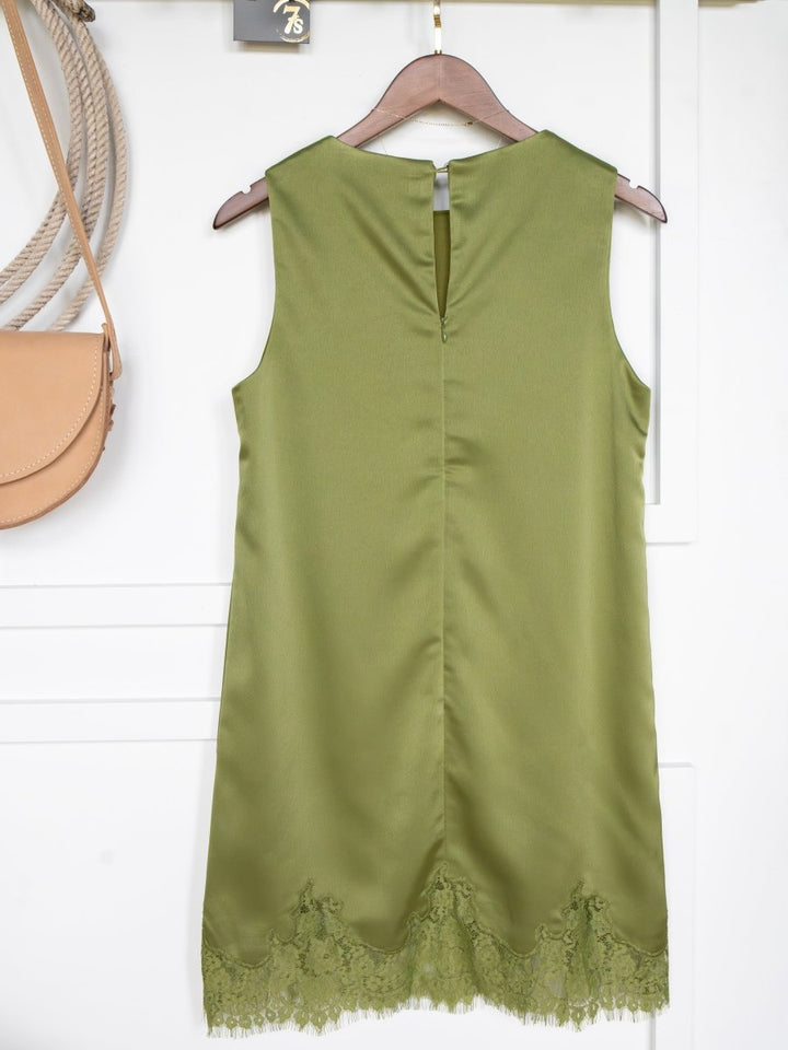 The Mesena Light Olive Satin Dress by Savannah Sevens western life{&}style, featuring a sleeveless cut, V-neckline, and lace hem, hangs on a wooden hanger against a white wall. A tan handbag is partially visible to the left.