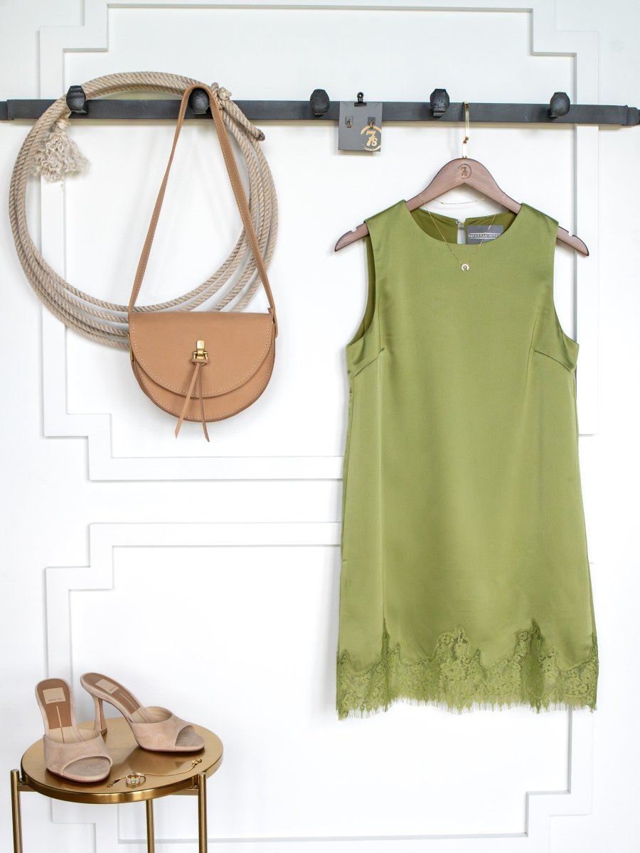 The Mesena Light Olive Satin Dress by Savannah Sevens western life{&}style hangs on the wall next to a tan crossbody bag, with tan heeled sandals placed on a gold side table against a white background.