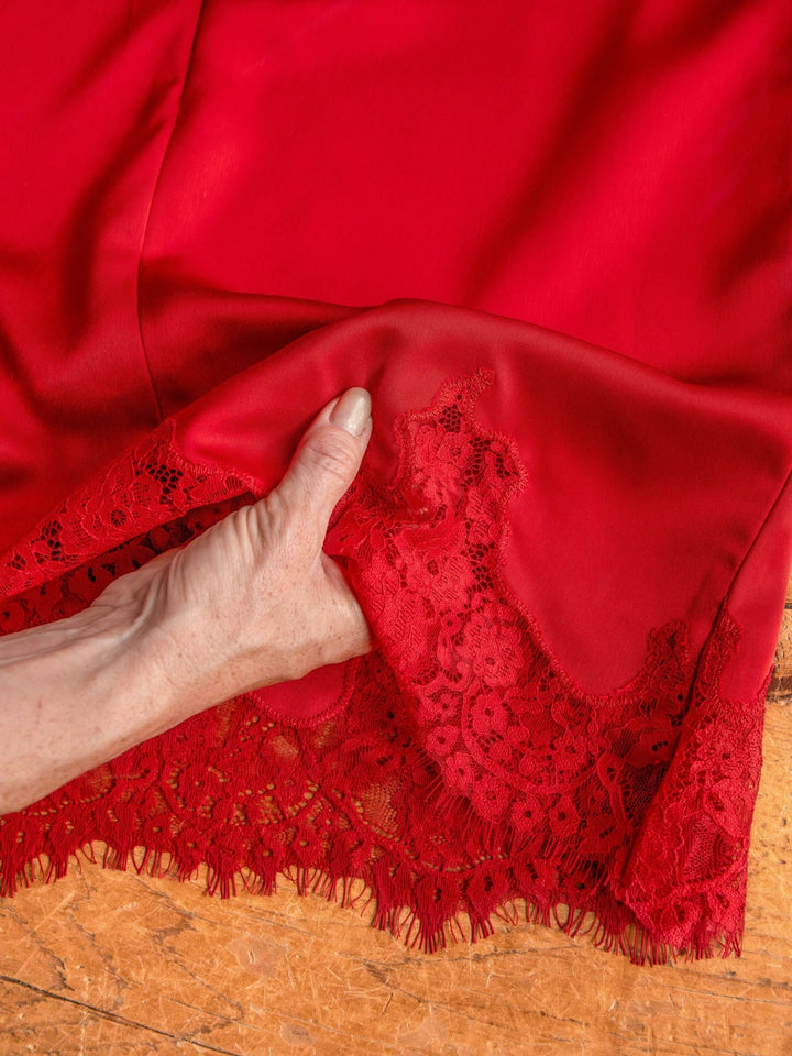 A hand lifts the hem of the Mesena Red Satin Dress by Savannah Sevens western life{&}style, revealing its delicate floral lace detail. The smooth, shiny satin stands out against a textured light brown background.