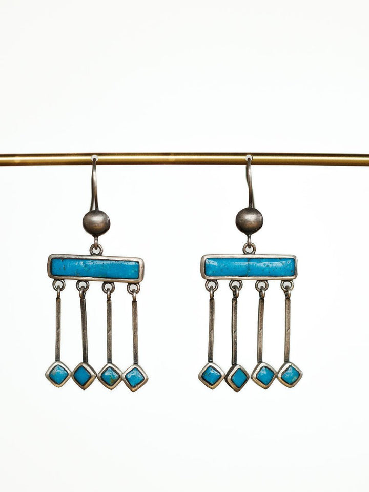 The Mesita Earrings by Savannah Sevens western life{&}style are handmade silver earrings with horizontal blue bars and three hanging rods, each set with a blue Kingman turquoise stone, shown on a gold rod against a white background.