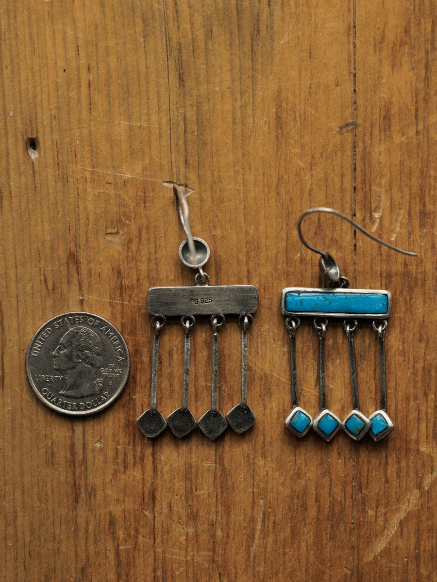Two Mesita Earrings by Savannah Sevens western life{&}style, featuring Kingman turquoise accents, rest on a wooden surface beside a US quarter for scale; one earring is missing stones and a hook, while the other is intact.