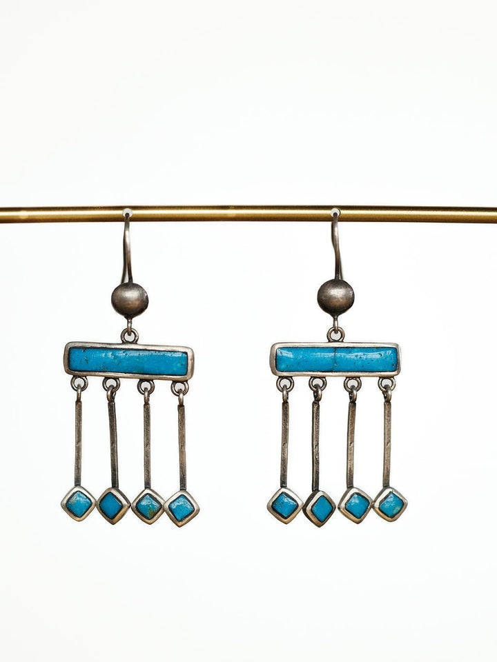 The Mesita Earrings by Savannah Sevens western life{&}style are handmade silver earrings with horizontal blue bars and three hanging rods, each set with a blue Kingman turquoise stone, shown on a gold rod against a white background.