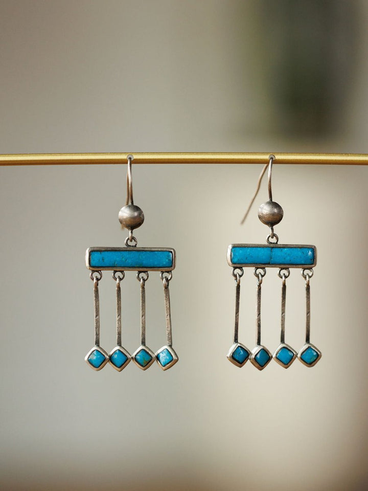 The Mesita Earrings by Savannah Sevens western life{&}style are handmade silver earrings with rectangular Kingman turquoise bars and three hanging silver strands tipped with small diamond-shaped turquoise stones, shown on a gold rod against a neutral background.