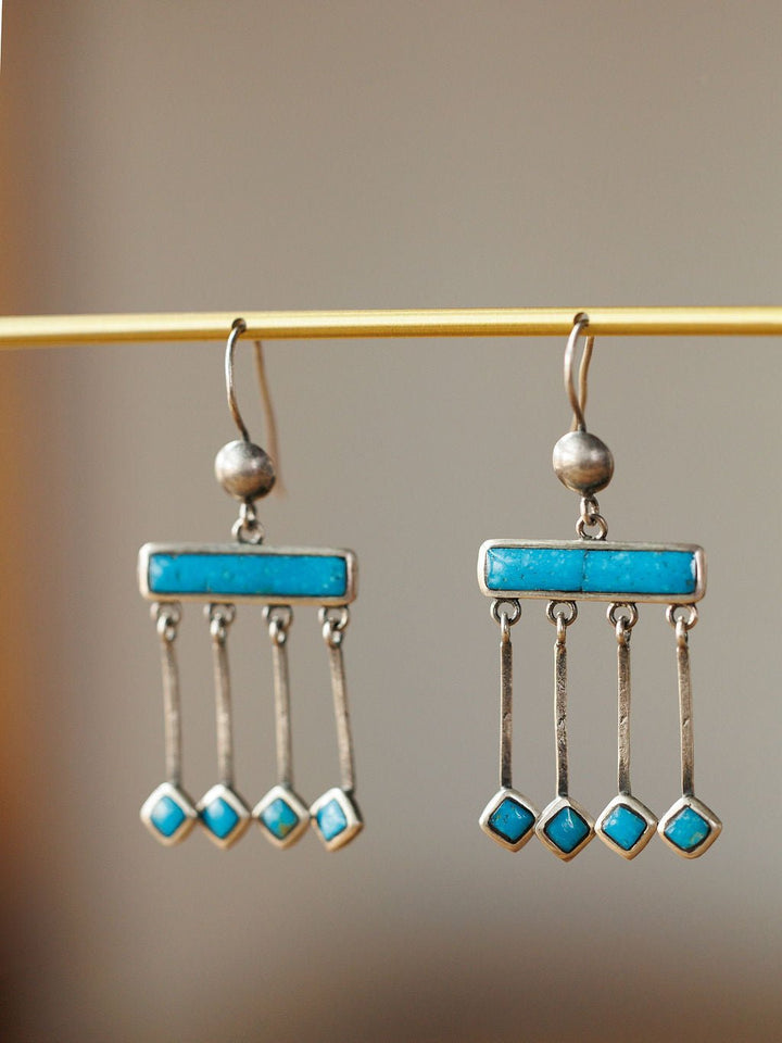 The Mesita Earrings by Savannah Sevens western life{&}style are handmade silver earrings, each featuring four rectangular Kingman turquoise stones, displayed on a horizontal rod with a soft blurred background.