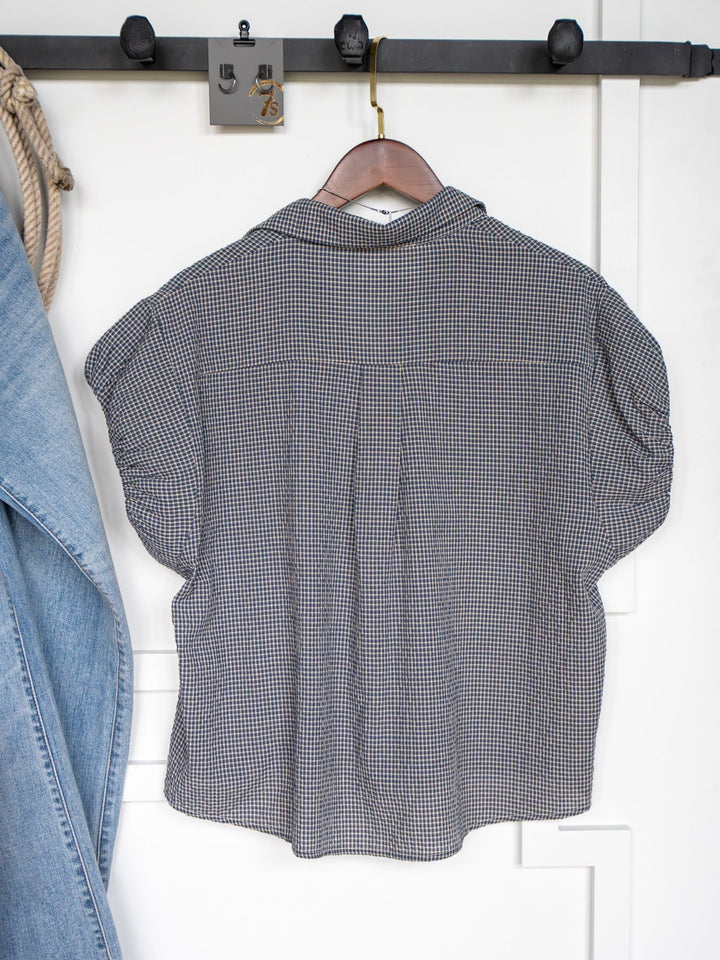 The Metairie plaid short sleeve top by Savannah Sevens western life{&}style, featuring a relaxed fit, hangs on a wooden hanger against a white wall with part of a blue denim jacket visible to the left.