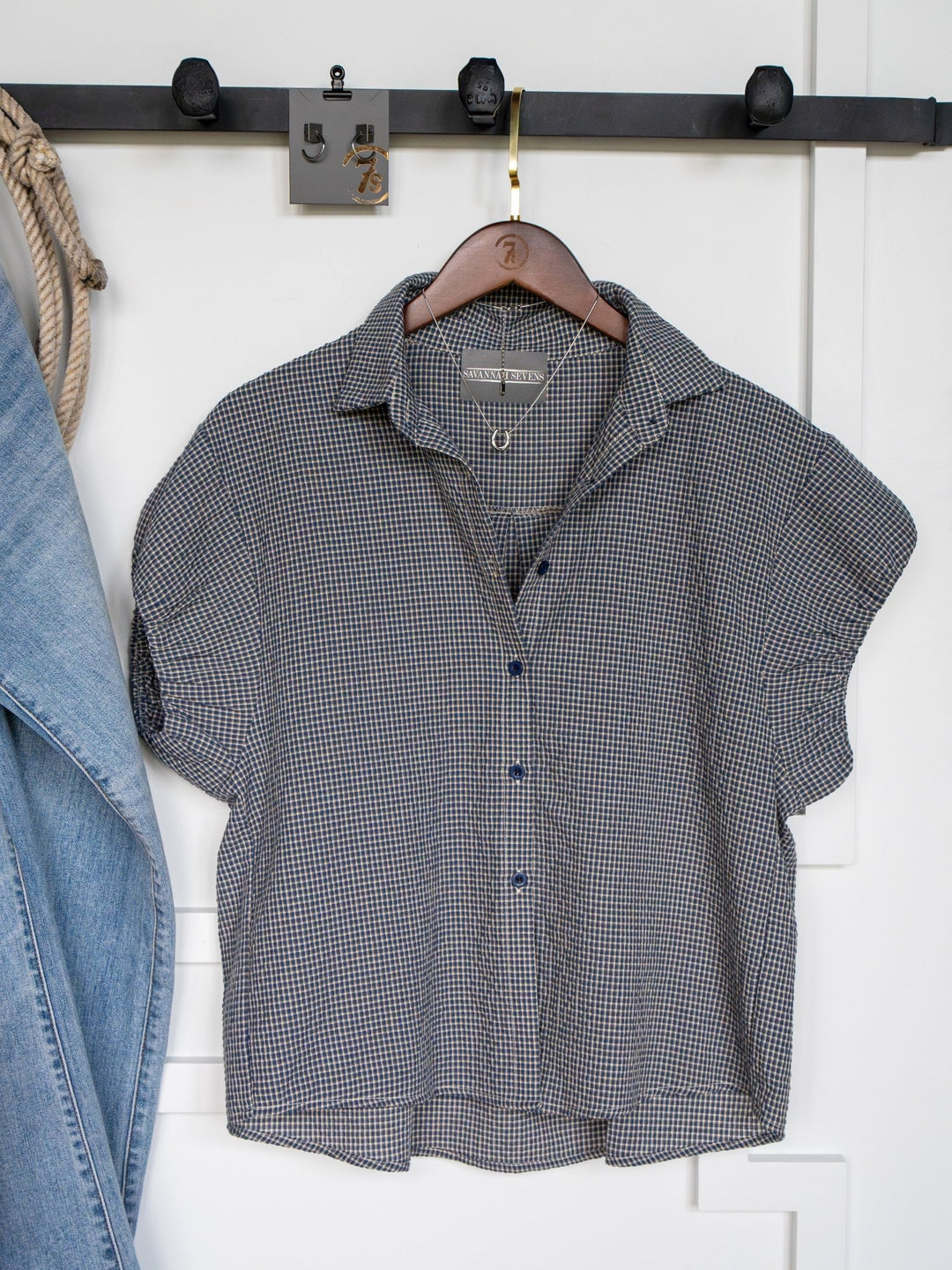The Metairie short sleeve plaid top by Savannah Sevens western life{&}style hangs on a wooden hanger from a black wall hook, with part of a denim jacket to the left and a white wall in the background.