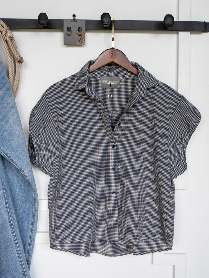 The Metairie short sleeve plaid top by Savannah Sevens western life{&}style hangs on a wooden hanger from a black wall hook, with part of a denim jacket to the left and a white wall in the background.