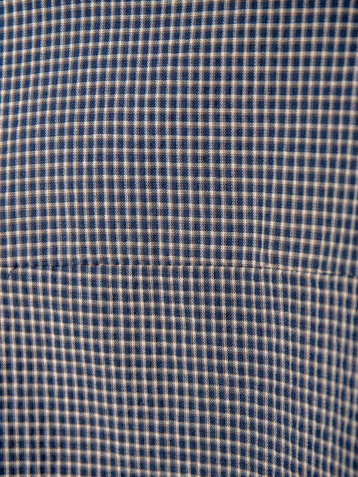 Close-up of The Metairie by Savannah Sevens western life{&}style, featuring a cotton blend fabric in a small blue and beige checkered pattern. Texture, stitching, and a horizontal seam highlight the relaxed fit of this plaid short sleeve top.