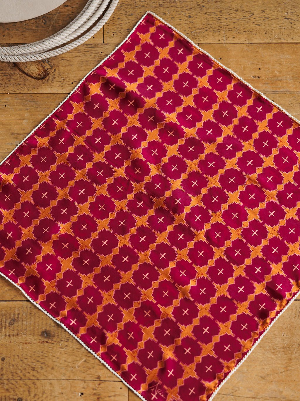 The Mettawa Neck Scarf by Savannah Sevens western life{&}style, featuring a red and orange geometric silk pattern with hand-drawn accents and a white border, is displayed flat on wood beside a coiled rope in the top left.