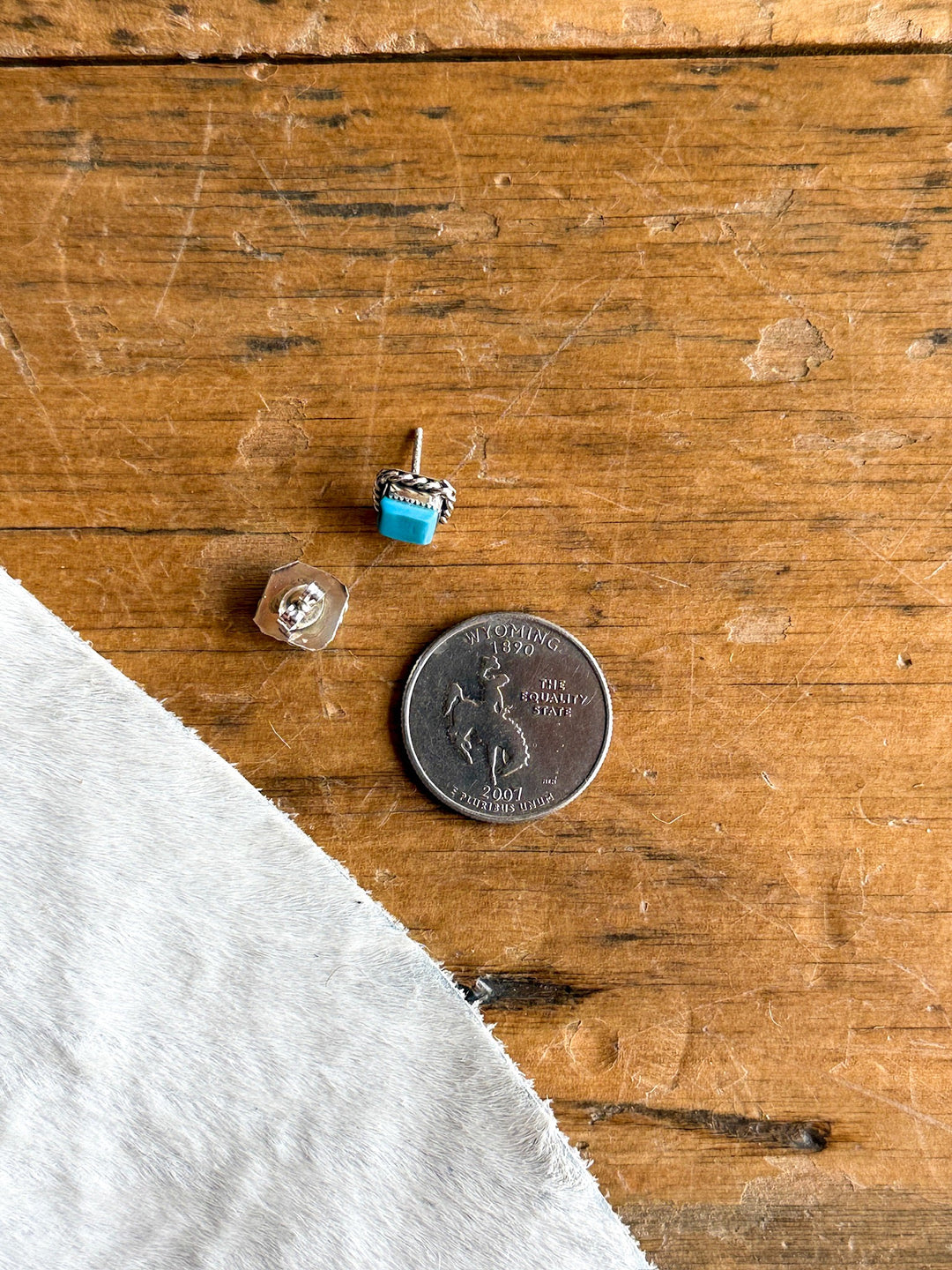 A pair of Mexia Turquoise Earrings from Savannah Sevens western life{&}style, a crystal stud earring, and a Wyoming state quarter rest on a wooden surface next to a piece of white fabric.
