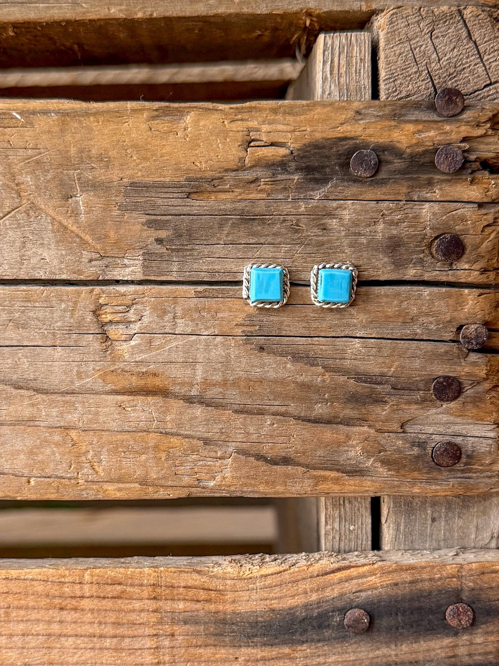 A pair of Mexia Turquoise Earrings by Savannah Sevens western life{&}style, featuring square Kingman turquoise stones with silver borders, are displayed on a rustic wooden surface with visible nails and grain patterns.