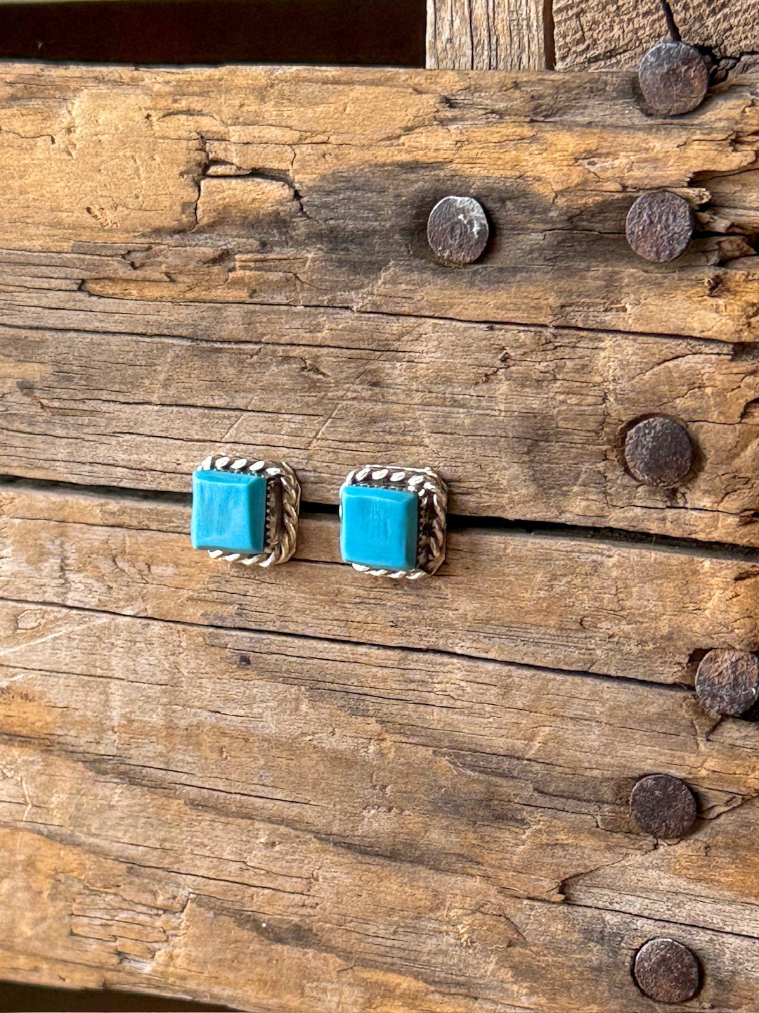 The Mexia Turquoise Earrings by Savannah Sevens western life{&}style, featuring square Kingman turquoise stones and intricate silver rope detailing, are showcased on a rustic wooden surface with visible nails and distinct grain patterns.
