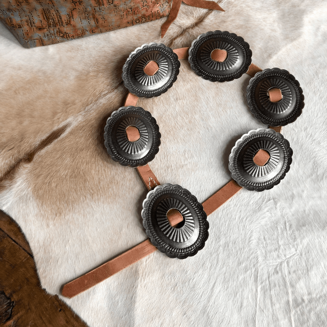 The Modesto Belt by Savannah Sevens western life{&}style features six large, oval silver conchos with scalloped edges and engraved details on distressed harness leather, displayed on a tan fur surface.