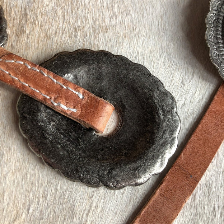 A close-up of a round, scalloped silver concho with a central slot, threaded with distressed harness leather. Set against a light, textured background. Perfect accent for the Savannah Sevens western life{&}style Modesto Belt.