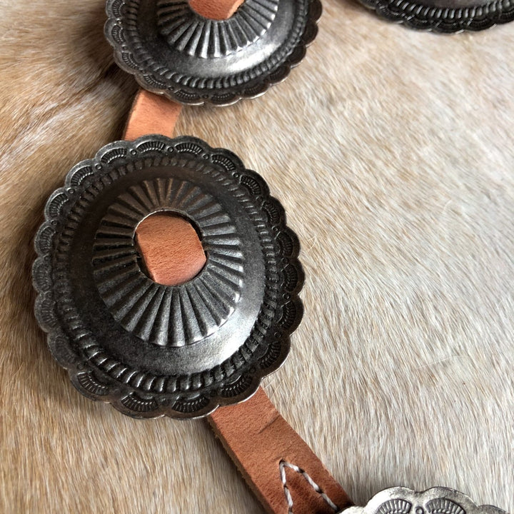 A close-up of the Modesto Belt by Savannah Sevens western life{&}style, featuring silver oval conchos with scalloped edges and intricate engravings on tan distressed harness leather, displayed on light brown fur.