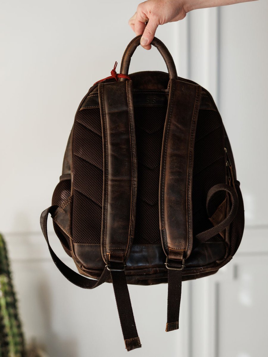 A hand holds up the Mohave Backpack by Savannah Sevens western life{&}style, showing its brown leather back and padded shoulder straps against a light, blurred background that highlights this concealed carry backpack’s classic design.