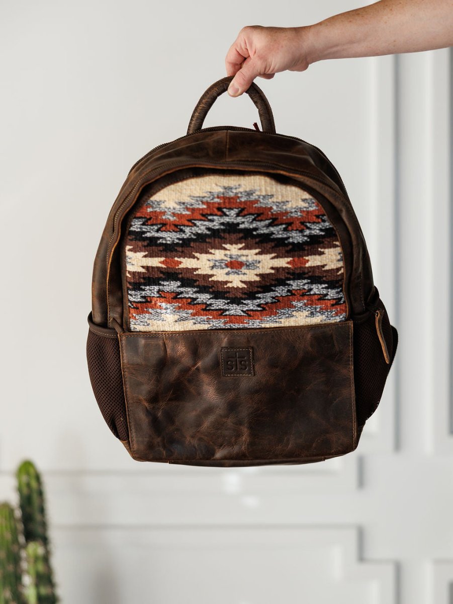 A hand holds up the Mohave Backpack by Savannah Sevens western life{&}style, a brown leather bag with a colorful southwestern woven front panel, mesh side pockets, and a rustic textured look inspired by classic saddle blanket designs.