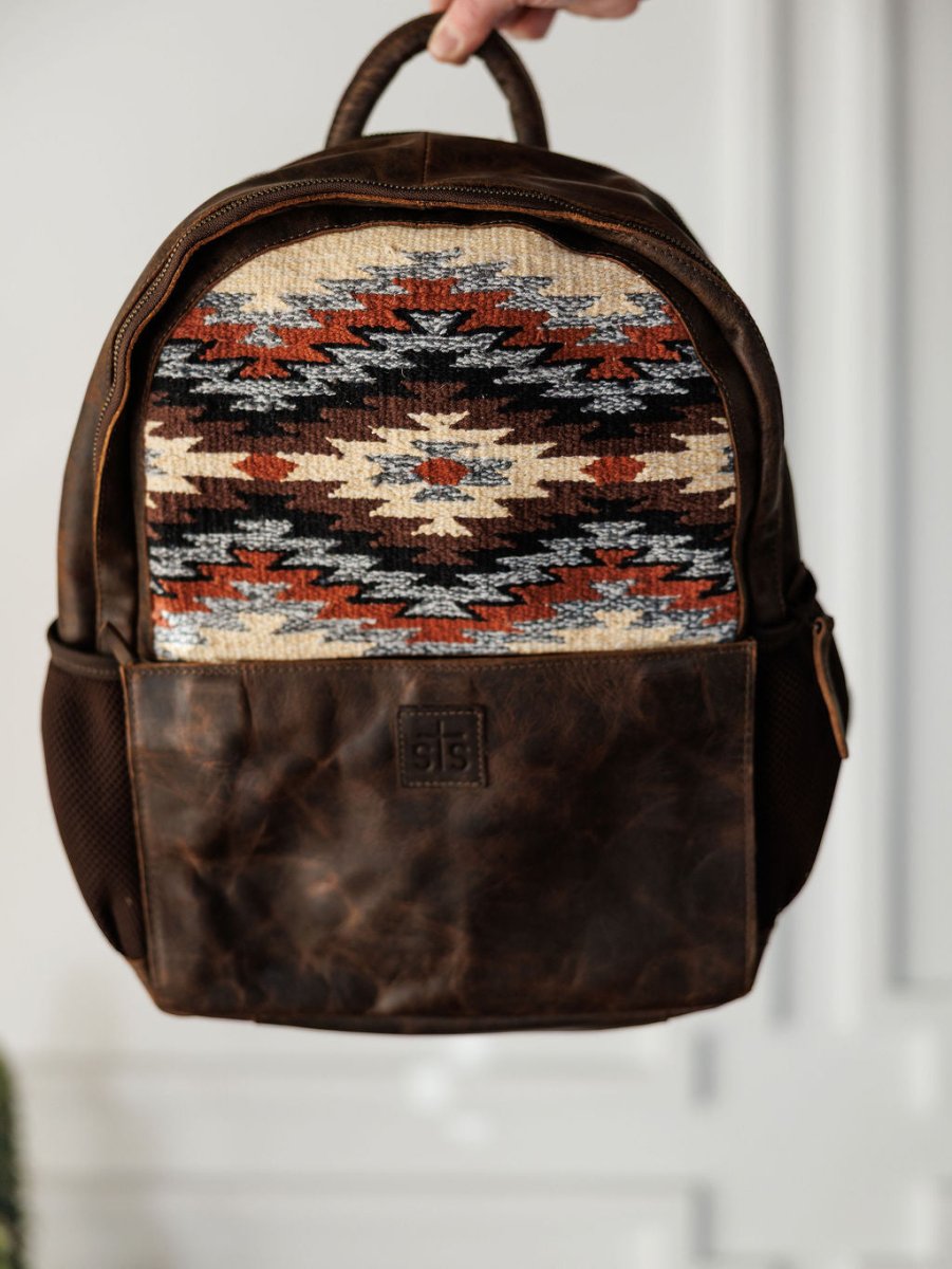 A hand holds the Mohave Backpack by Savannah Sevens western life{&}style, featuring a brown leather saddle blanket design with a colorful Southwestern geometric pattern, front pocket, and short top handle.