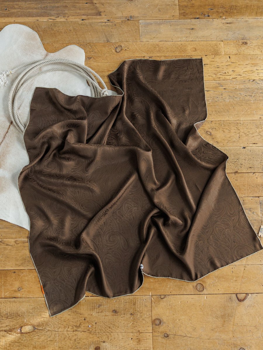 The Mokane Wild Rag by Savannah Sevens western life{&}style, a brown paisley jacquard silk scarf, is draped on a wooden floor beside a white cowboy hat and a coiled rope.