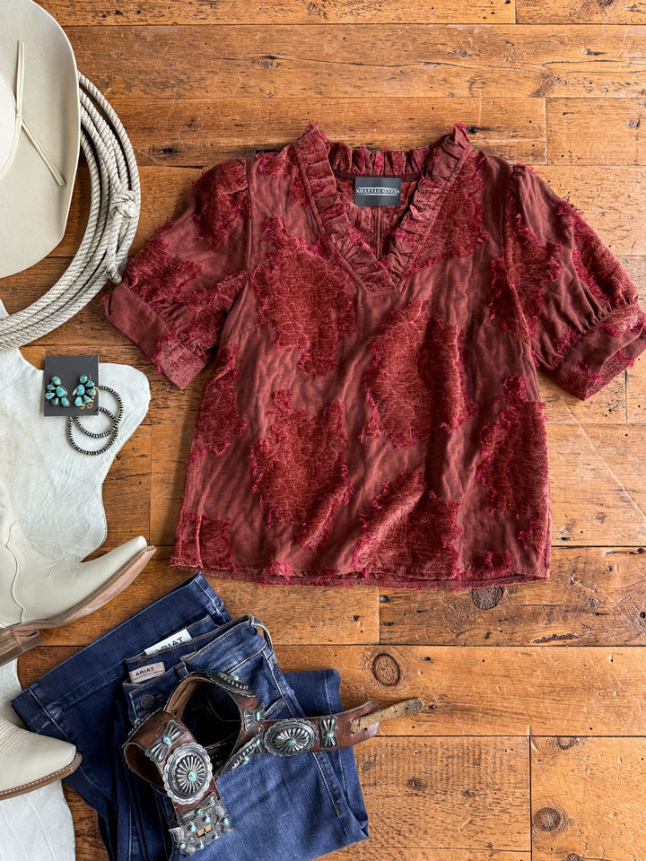 The Mokena Berry Textured Top by Savannah Sevens western life{&}style, featuring a rich red hue with a velvet floral pattern, is displayed on a wooden floor alongside a white cowboy hat, turquoise earrings, white boots, blue jeans, and a leather belt with an ornate buckle.