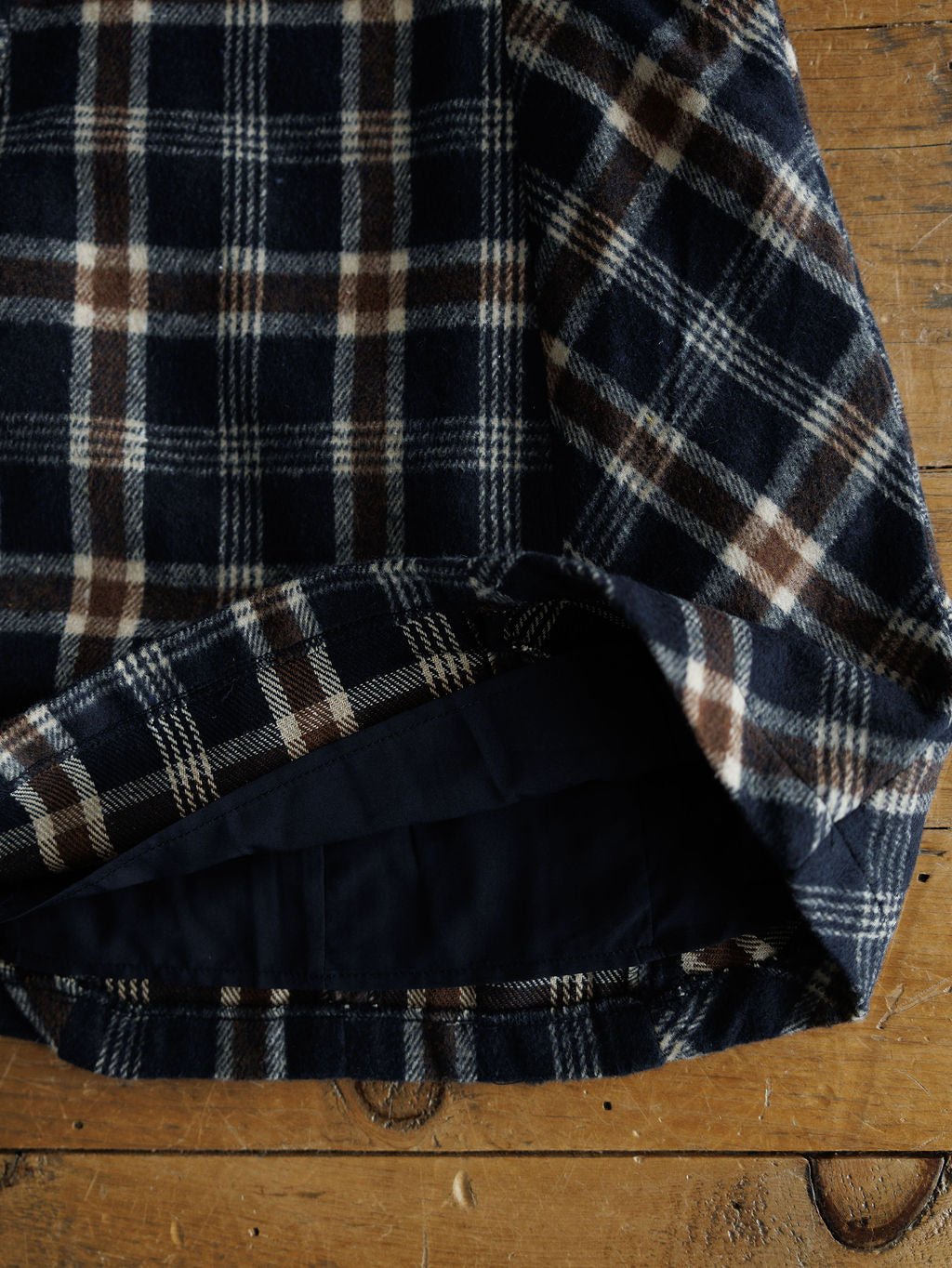A close-up of the Mondovi Flannel Dress by Savannah Sevens western life{&}style, featuring navy blue, brown, and white plaid with a folded hem resting on a wooden surface.
