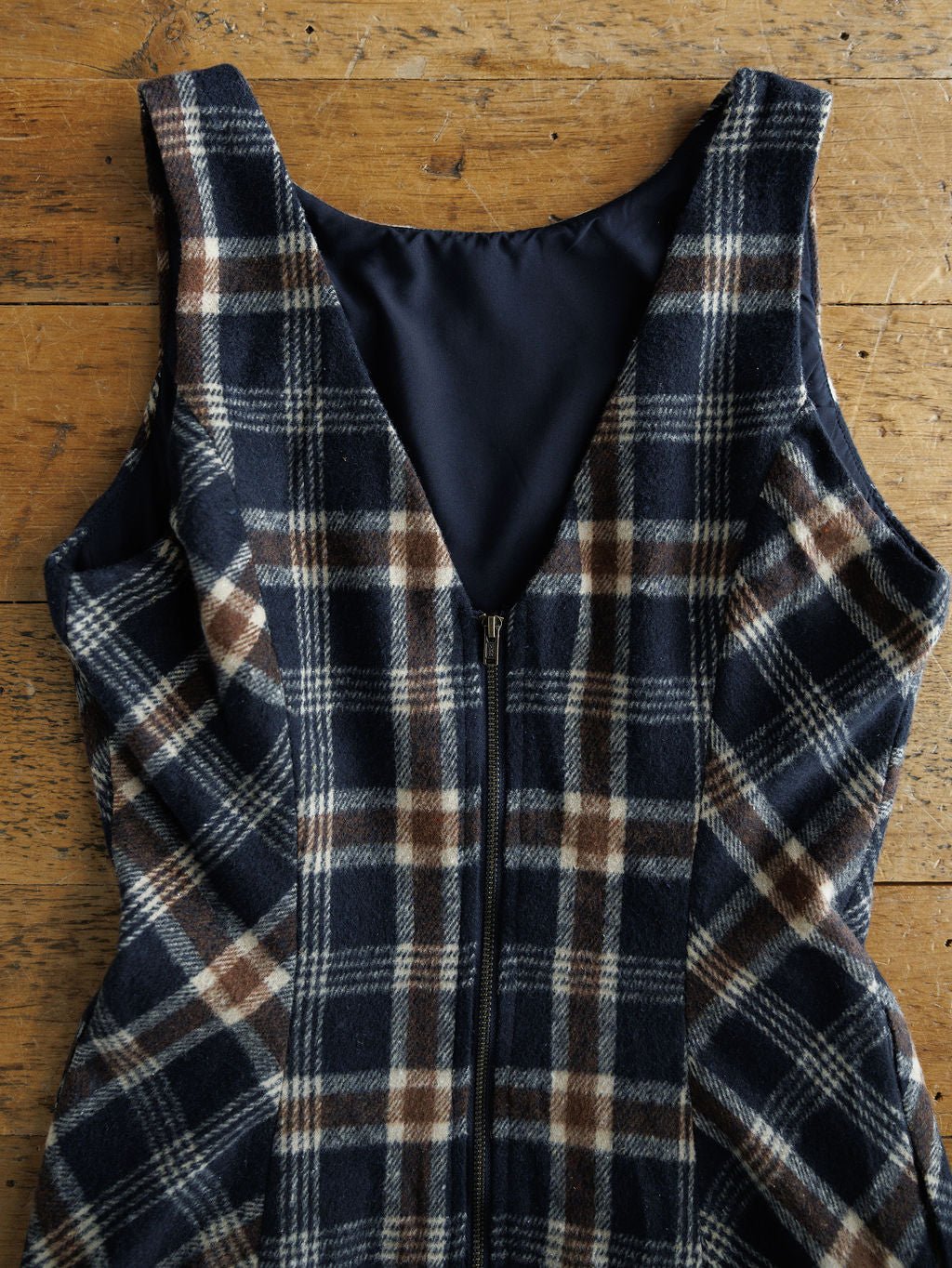The Mondovi Flannel Dress by Savannah Sevens western life{&}style, a sleeveless plaid dress with a deep V-neck and zipper front in navy, brown, and white checks, is displayed flat on a rustic wooden surface.