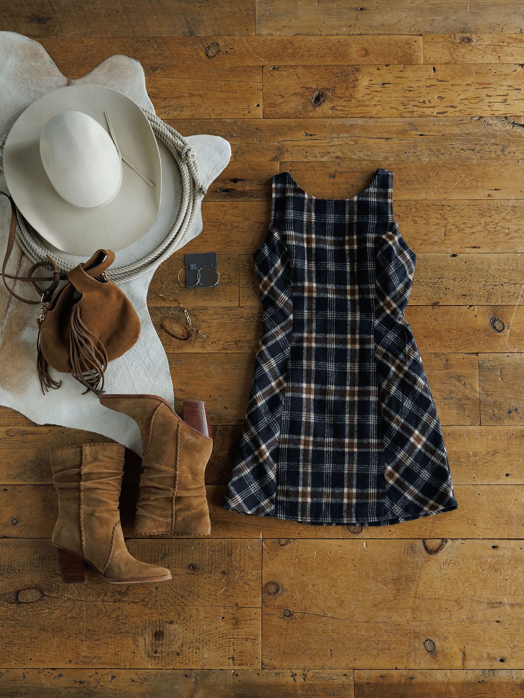 The Mondovi Flannel Dress by Savannah Sevens western life{&}style is styled with tan suede boots, a wide-brimmed hat, fringed suede bag, brown belt, and silver bracelet on a wooden floor atop a white cowhide rug.