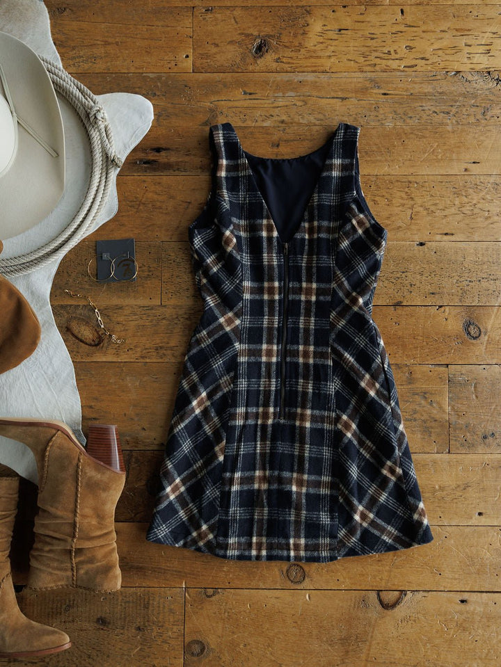 The Mondovi Flannel Dress by Savannah Sevens western life{&}style is laid flat on a wooden floor, styled with a white hat, suede heeled boots, and silver hoops—a chic young contemporary dress ensemble.