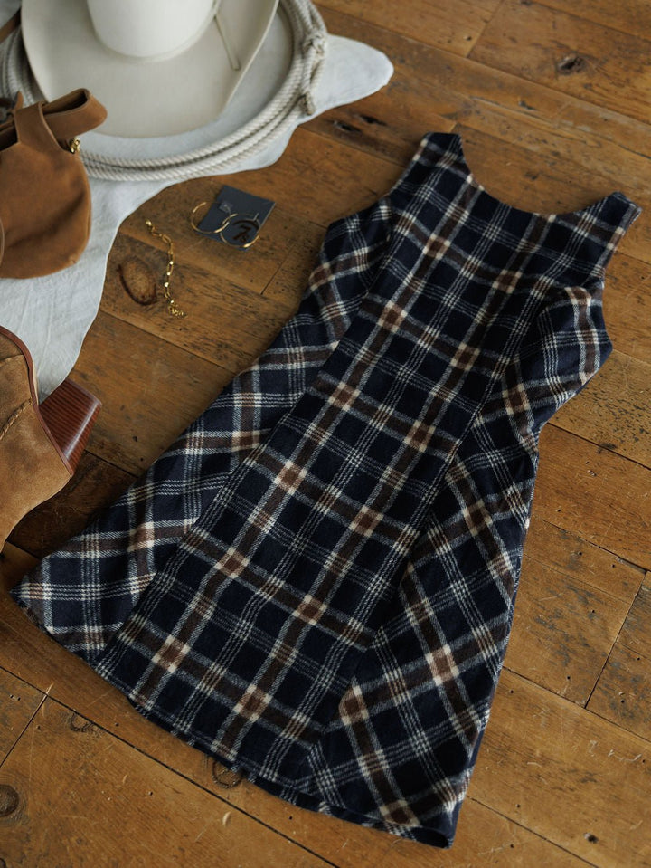The Mondovi Flannel Dress by Savannah Sevens western life{&}style, a sleeveless plaid dress in navy, brown, and white, is styled flat with brown boots, a beige hat, gold jewelry, and earrings for versatile young contemporary looks.