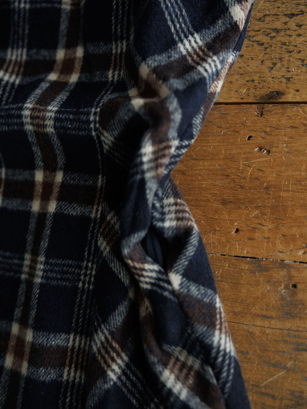 A close-up of the Mondovi Flannel Dress by Savannah Sevens western life{&}style, featuring navy blue, brown, and white plaid fabric draped with gentle wrinkles on a worn wooden surface.