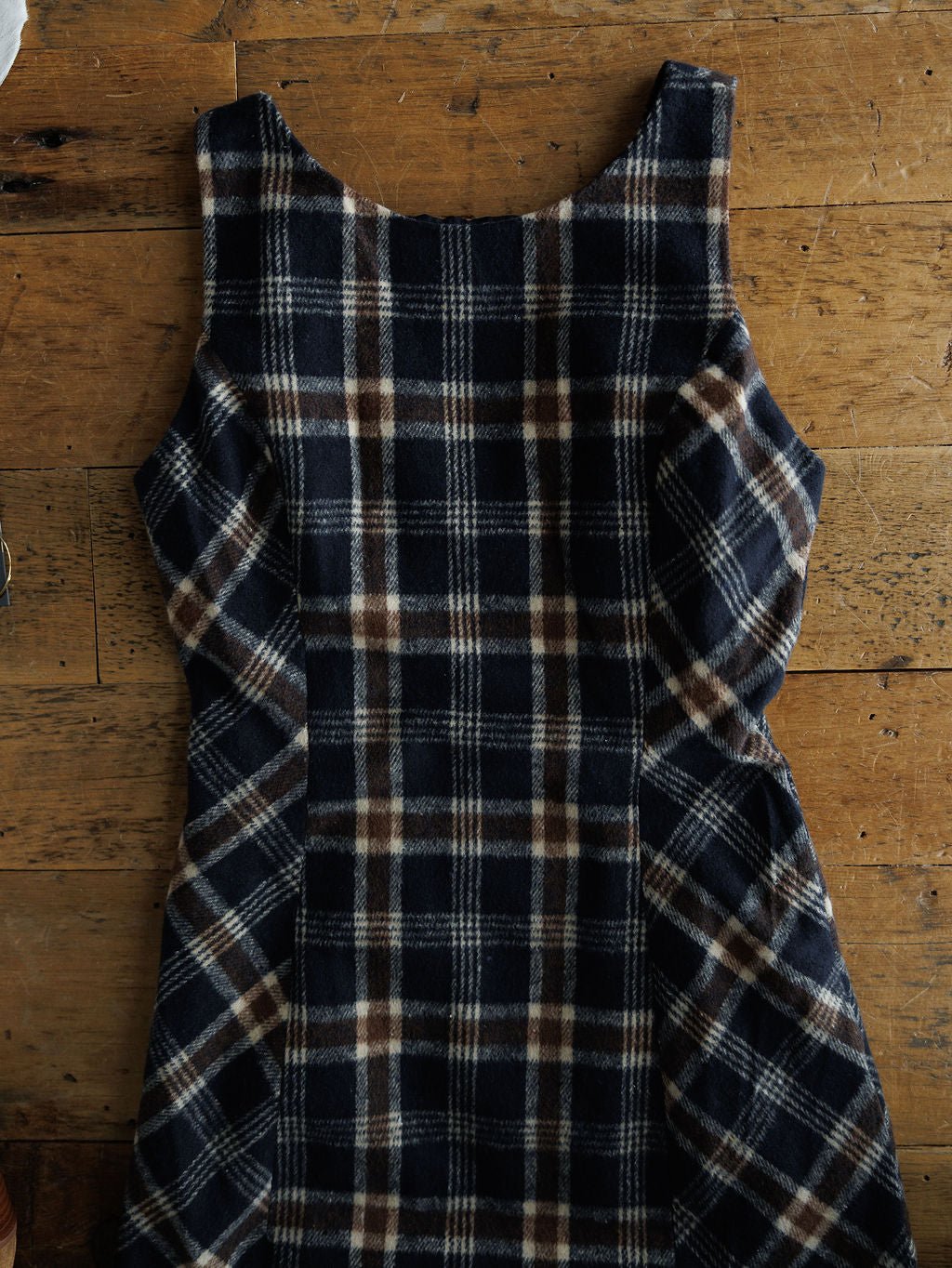 The Mondovi Flannel Dress by Savannah Sevens western life{&}style, a young contemporary sleeveless dress with a blue, brown, and white plaid pattern, is laid flat on a wooden surface.
