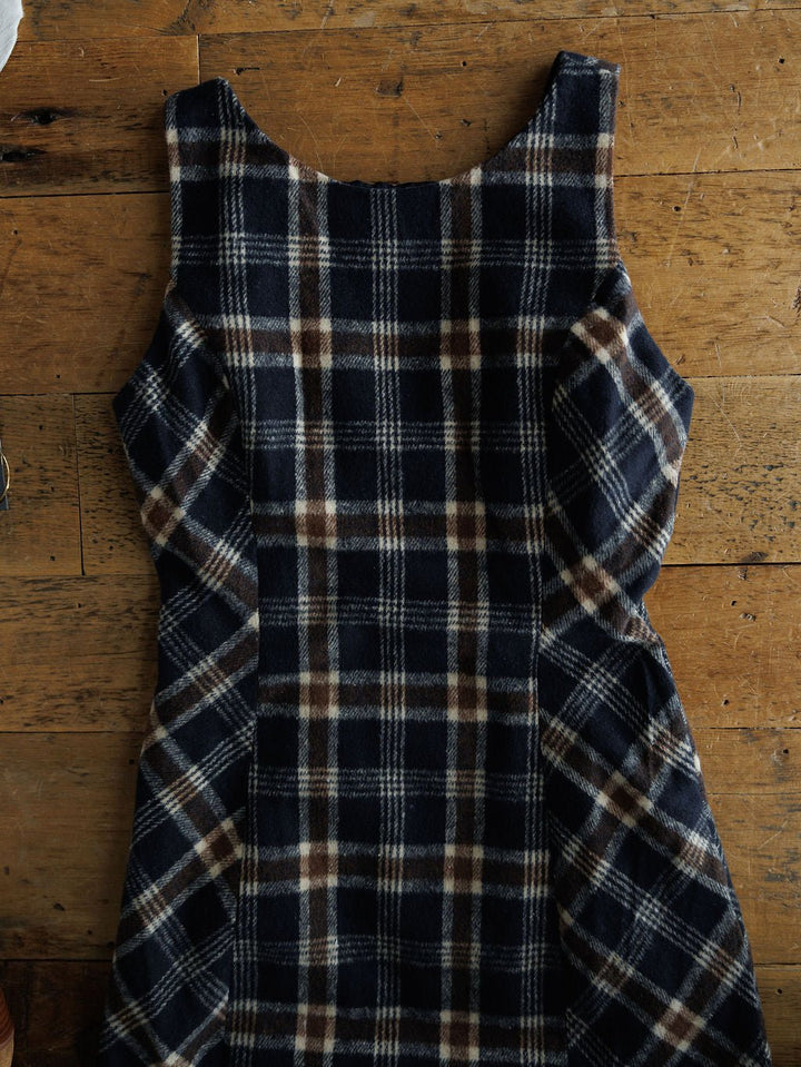 The Mondovi Flannel Dress by Savannah Sevens western life{&}style, a young contemporary sleeveless dress with a blue, brown, and white plaid pattern, is laid flat on a wooden surface.