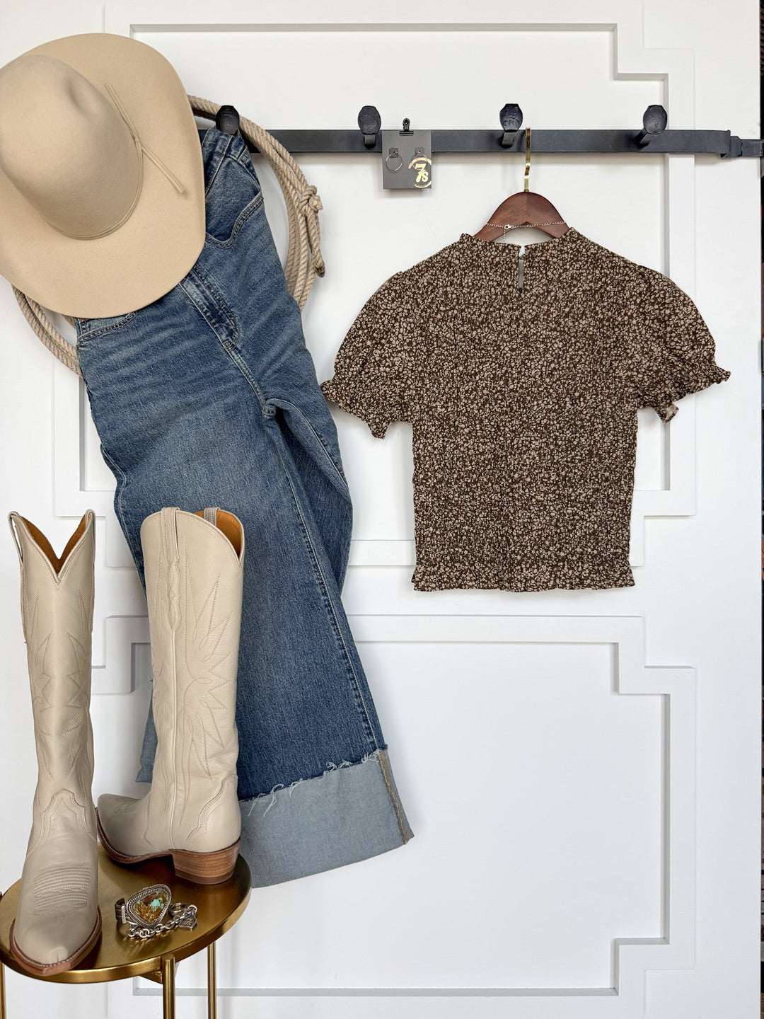 The Monrovia Brown Floral Top by Savannah Sevens western life{&}style is styled with a cowboy hat, cuffed blue jeans, and cream cowboy boots, all arranged against a white wall. Accessories—such as rings and a belt—are neatly displayed on a small dish nearby.