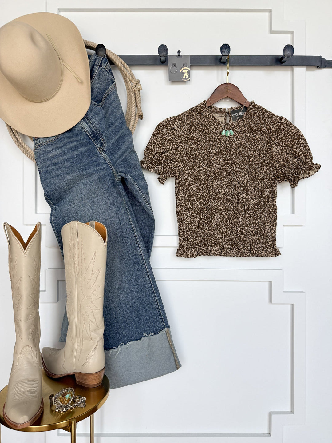 The Monrovia Brown Floral Top by Savannah Sevens western life{&}style is styled with a beige cowboy hat, cuffed blue jeans, and cream cowboy boots, all arranged against a white wall. Turquoise jewelry and a gold ring are displayed as accessories on a small table.