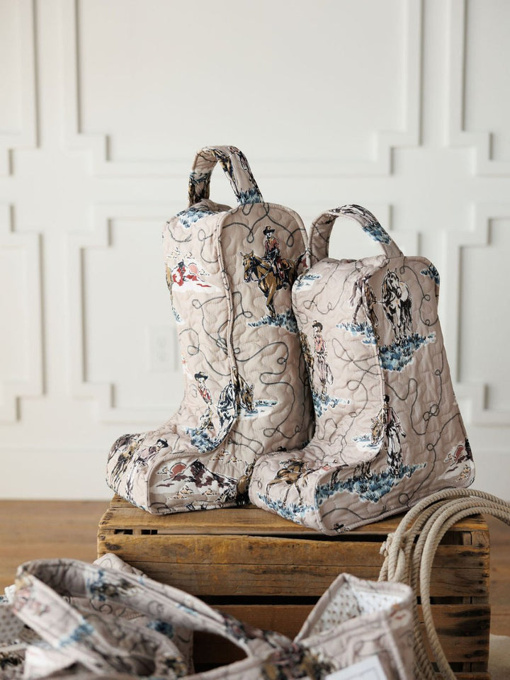 Two quilted boot bags from the Montana Summer Collection by Savannah Sevens western life&style, with Western-themed prints and large handles, sit on a wooden crate in a bright room. Nearby are a coiled rope and matching bag.