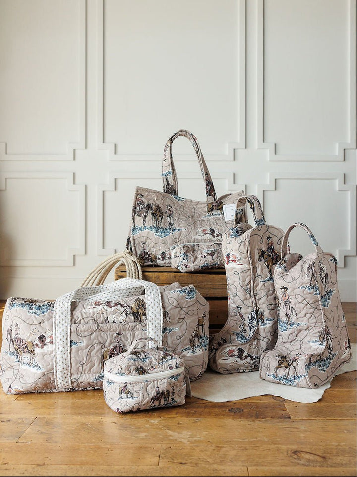 Five quilted travel bags and boots with a vintage map print from the Montana Summer Collection by Savannah Sevens western life&style are displayed on a wooden floor in front of a white paneled wall.