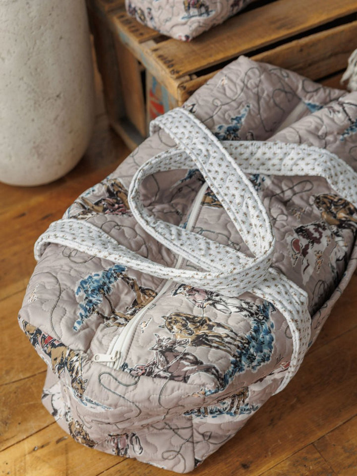 The Montana Summer Collection by Savannah Sevens western life&style includes a light gray quilted duffel with blue and brown floral prints, white zipper, patterned straps, and a matching small bag—both styled on wood surfaces.