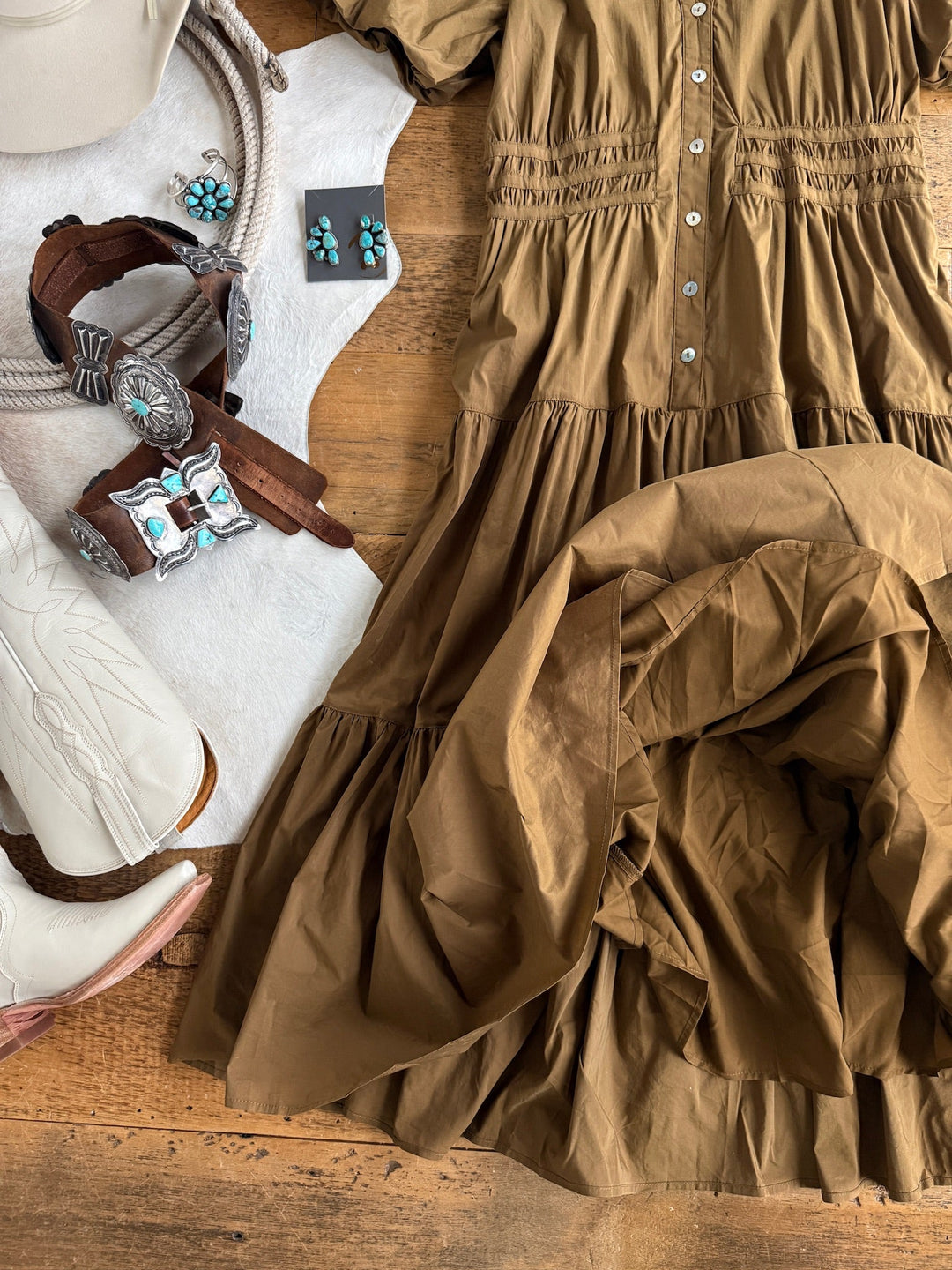 Flat lay featuring the Monterey Bronze Olive Puff Sleeve Dress by Savannah Sevens western life{&}style, paired with white cowboy boots, silver and turquoise jewelry, a beige hat, and a white rug displayed on a wooden surface.